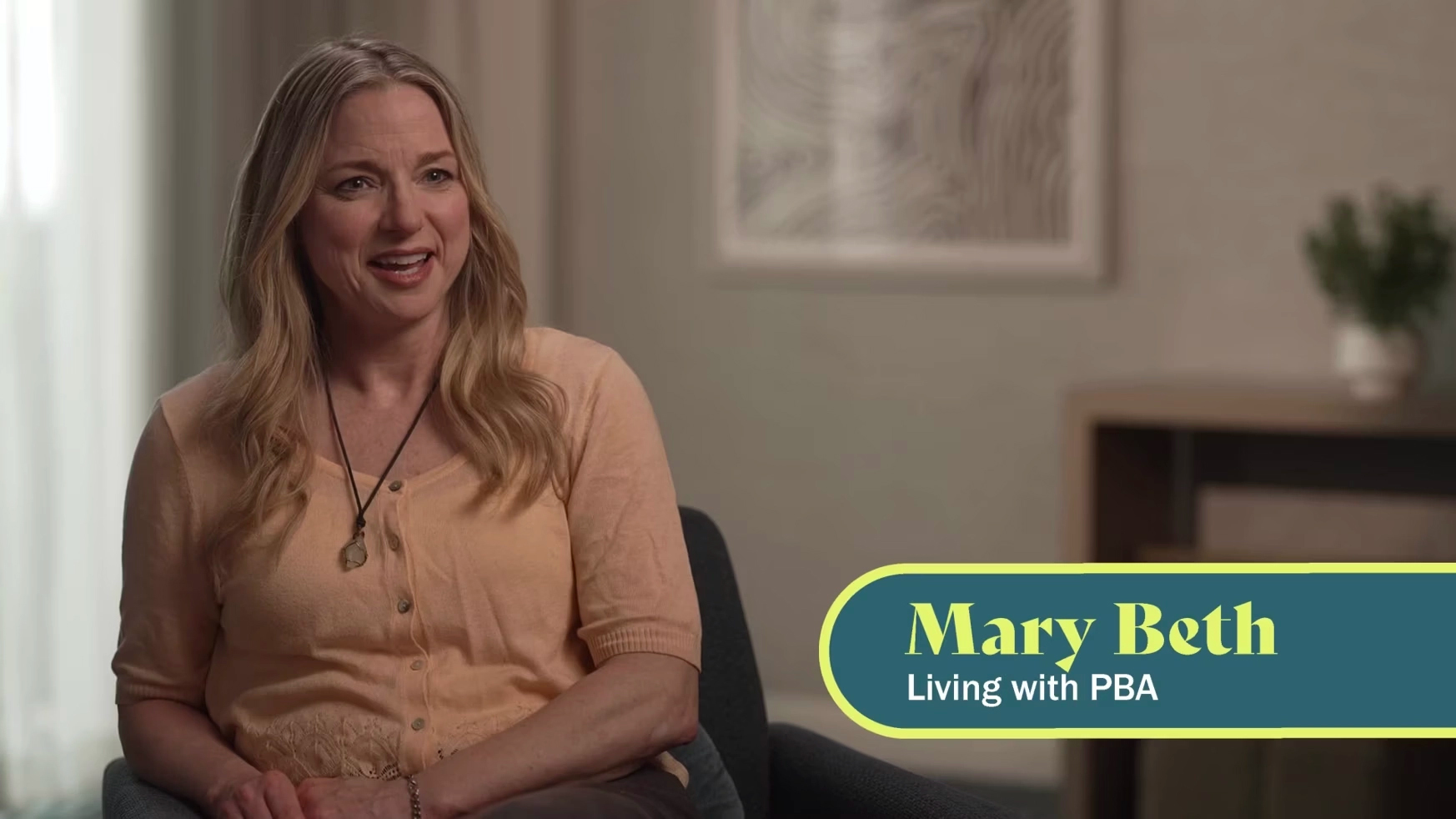 A white woman with long blonde hair is seen from the waist up, facing the camera. The caption on screen says “Mary Beth, living with PBA.”